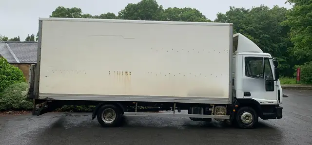 Side view of a long white box lorry used for removals, parked beside trees